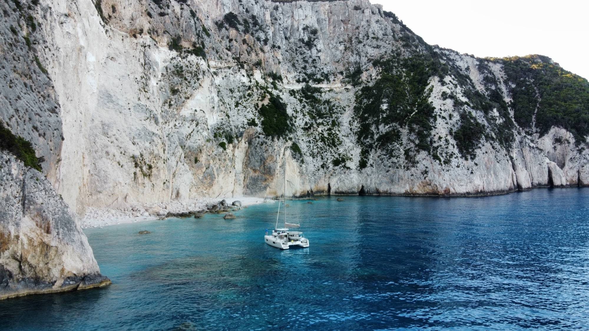 Ein Katamaran ankert in türkisem Wasser vor hohen weißen Kalkfelsen, dahinter ein schmaler Kiesstrand.