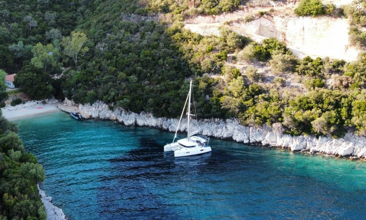 Ein Katamaran liegt in einer türkisen Bucht vor einem kleinen Strand, dahinter bewaldete Hänge.