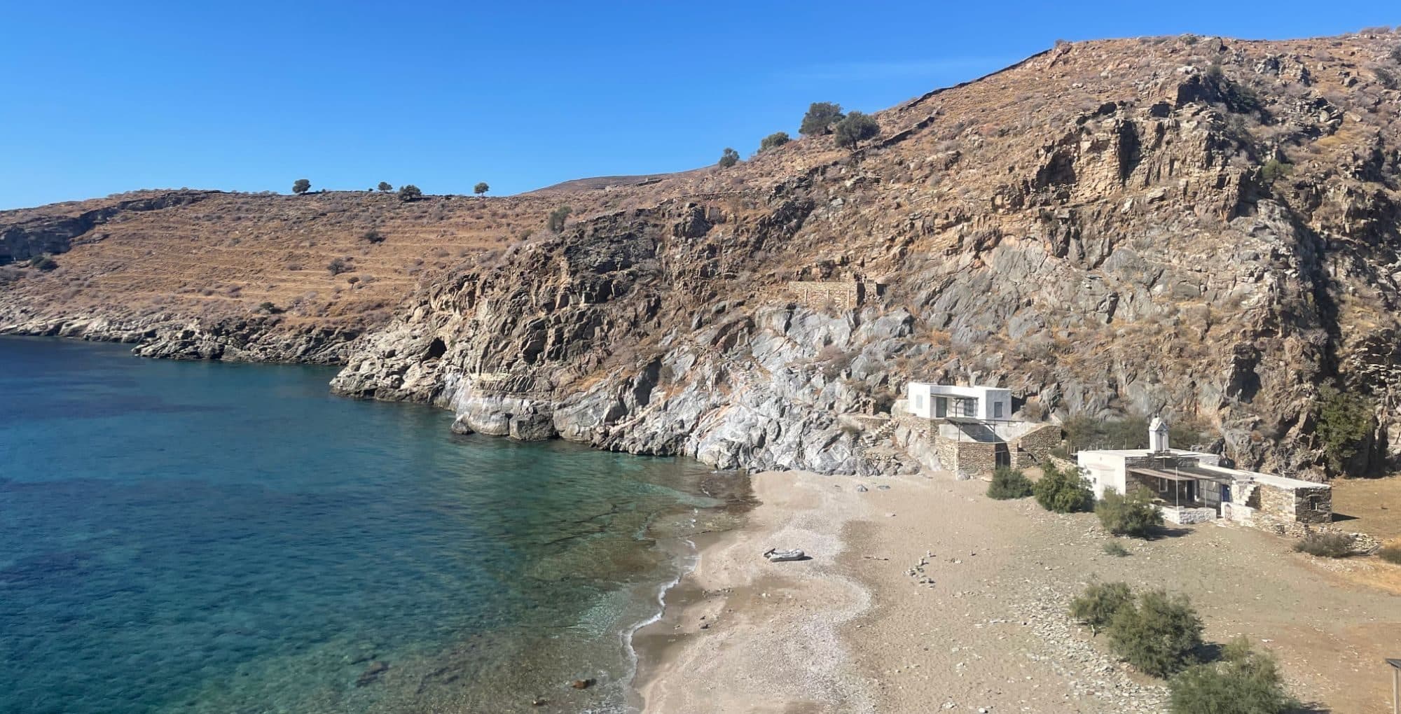 Ein Sandstrand in einer Bucht mit klarem Wasser, am Ufer ein Beiboot und zwei weiße Häuser unter einem kargen Hang.